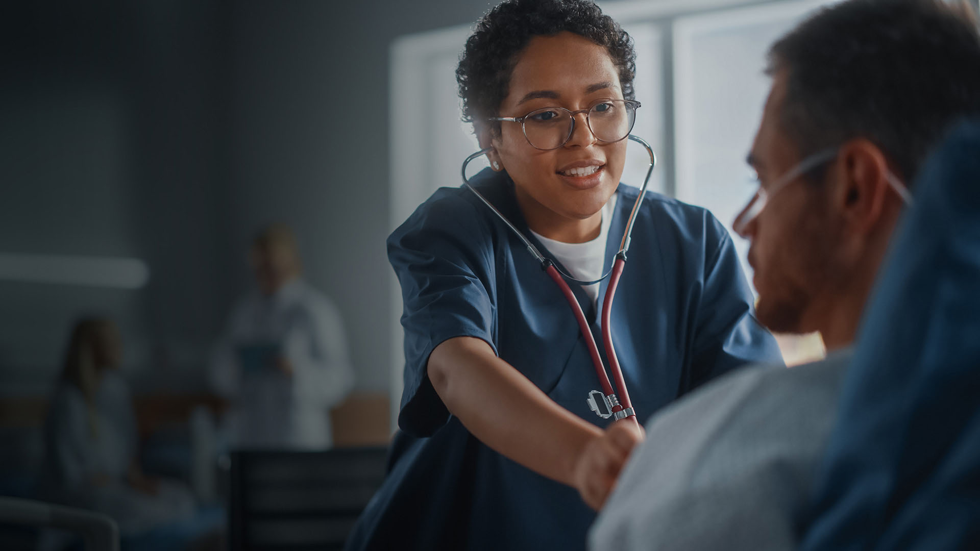 Hospital Ward: Friendly Black Head Nurse Uses Stethoscope to Listen to Heartbeat and Lungs of Recovering Male Patient Resting in Bed, Does Checkup. Man Getting well after Successful Surgery