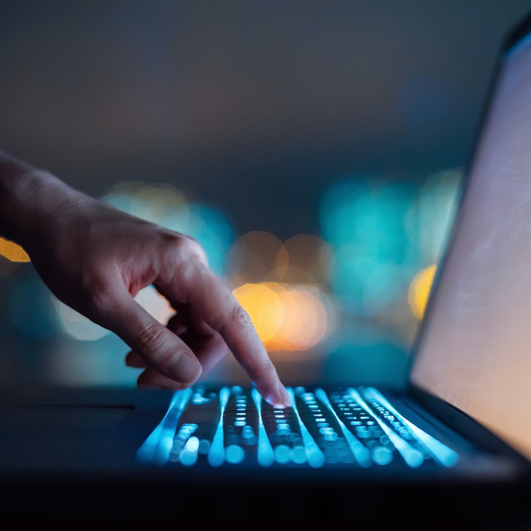 A person is engaging with a glowing laptop keyboard in a dimly lit environment, surrounded by a blurred cityscape, focusing intently on their task in the evening.
