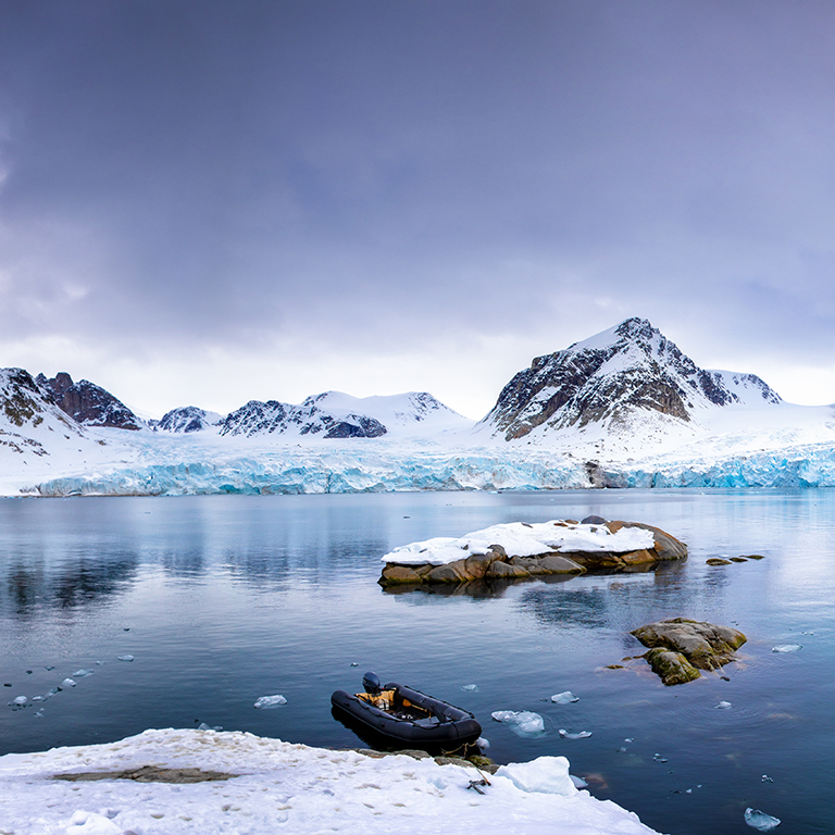 Panarama of the mountains, snow and blue glacial ice of the Smeerenburg glacier, Svalbard, and archipelago between mainland Norway and the North Pole. An inflatable boat is anchored in the foreground. 
