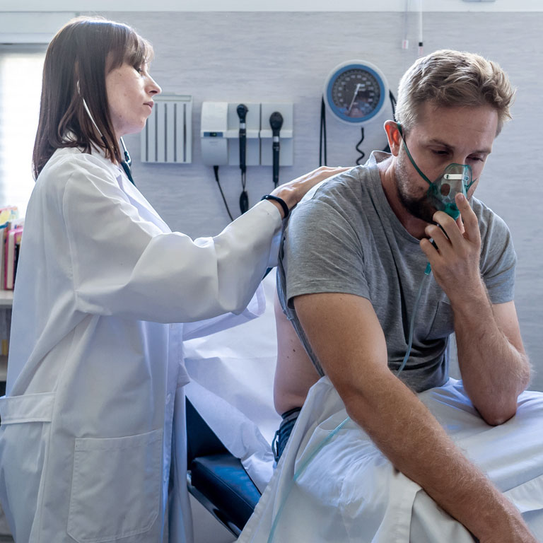 Young sick male patient with oxygen mask while female doctor listens his chest for respiratory disease with stethoscope in hospital emergency room