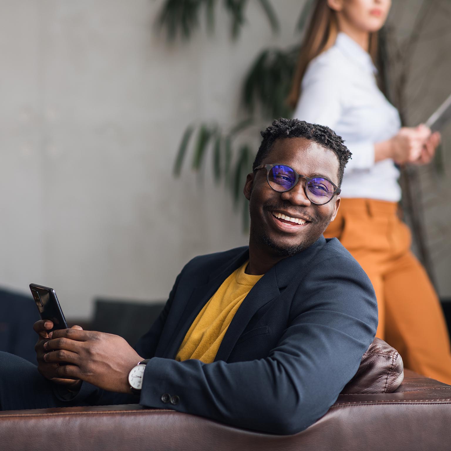 Happy young black man using a phone. Colleagues in the background. Joyful African-American businessman in a suit, sitting in a lobby, holding a phone.